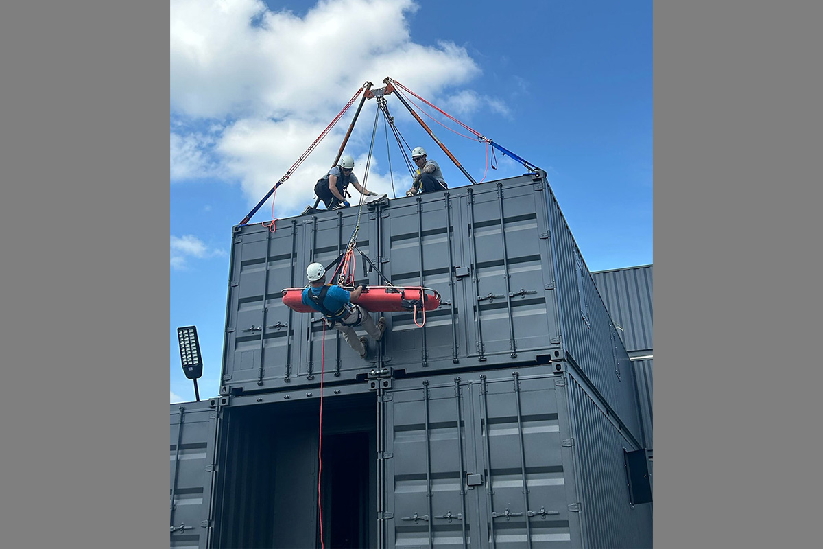 Large industrial container Tower being hoisted by a crane with red straps, as two firefighters in safety gear secure the load