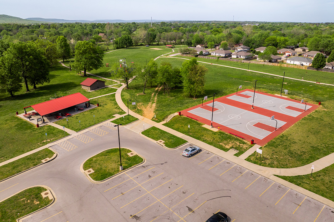 Aerial view of Farmington park featuring a large parking lot, a pavilion with a red roof, multiple basketball courts with bright red and gray surfaces, walking paths, and green fields surrounded by trees