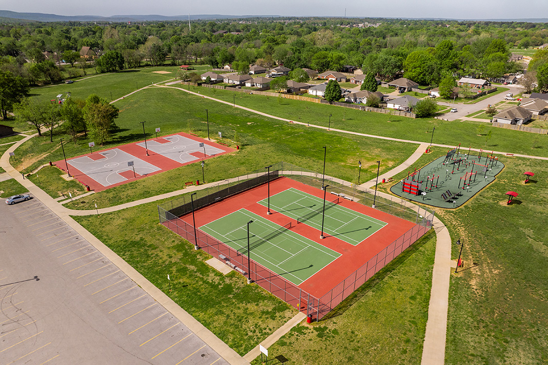 Aerial view of a community park featuring two red tennis courts with green outlines and two basketball courts, adjacent to a colorful playground area with various equipment, surrounded by a walking path and green grass.