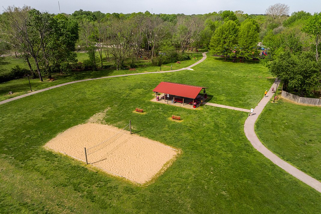 Aerial view of Farmington park with lush green grass and various amenities. Large sand volleyball court with a net set up in the middle, red-roofed pavilion with picnic tablese is a red-roofed pavilion with picnic tables underneath. 