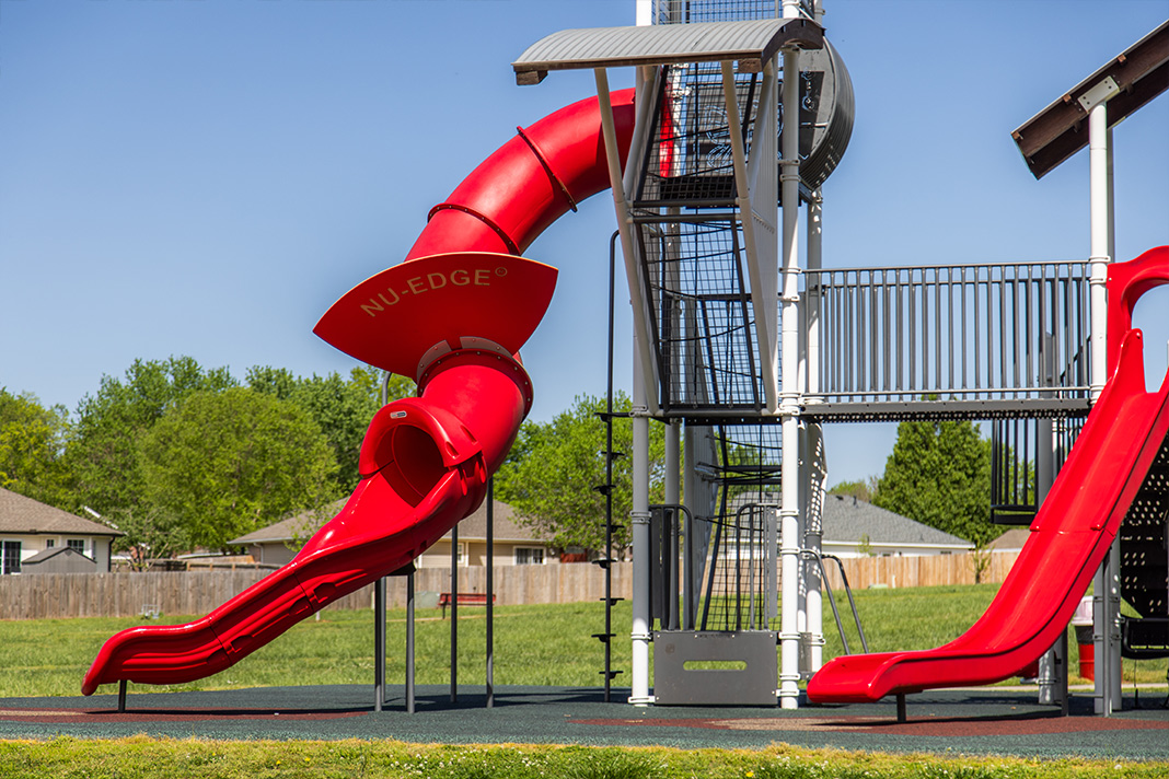 Close-up view of a modern playground in Farmington park featuring a large red spiral slide labeled 'NU-EDGE' alongside other climbing structures and slides