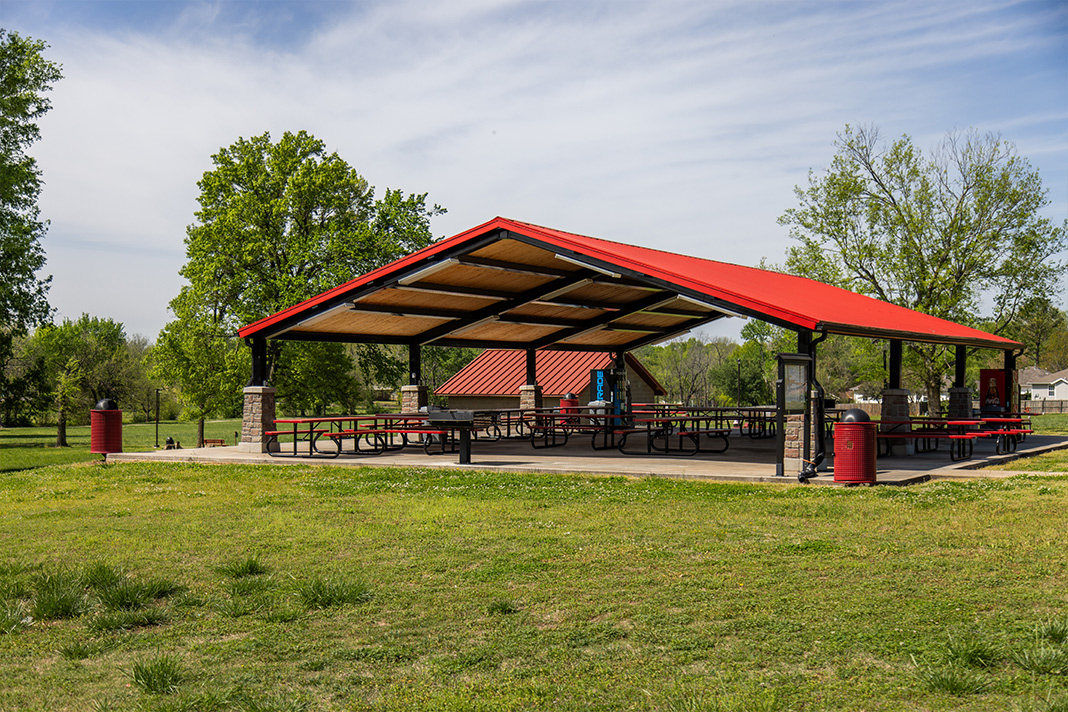 An outdoor park pavilion with a bright red roof and open sides, featuring numerous picnic tables under the shelter. The pavilion is surrounded by green grass and trees