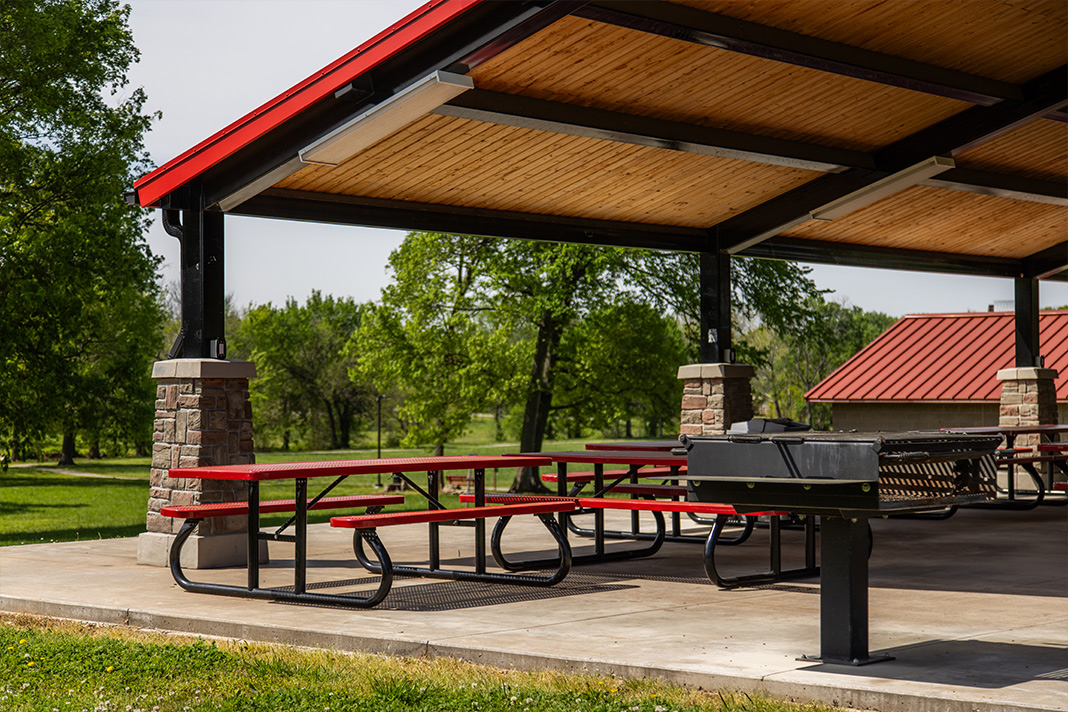 An outdoor park pavilion in Farmington park, featuring numerous picnic tables and bbq zone under the shelter. The pavilion is surrounded by green grass and trees