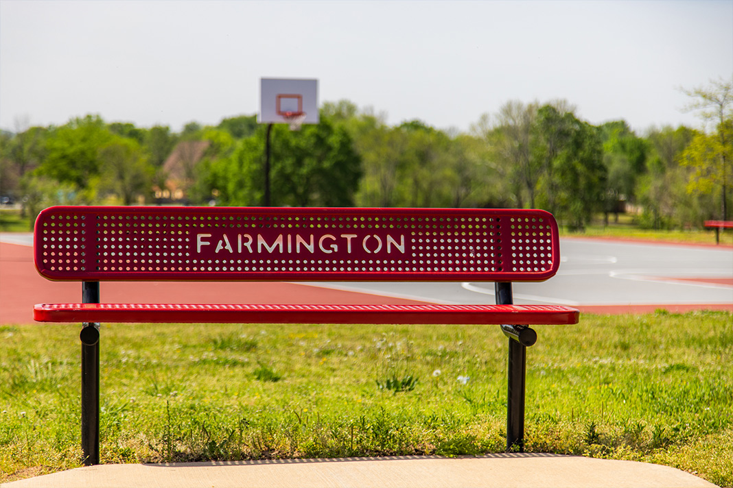 Red bench with the word 'Farmington' cut out of the backrest, situated in front of an outdoor basketball court