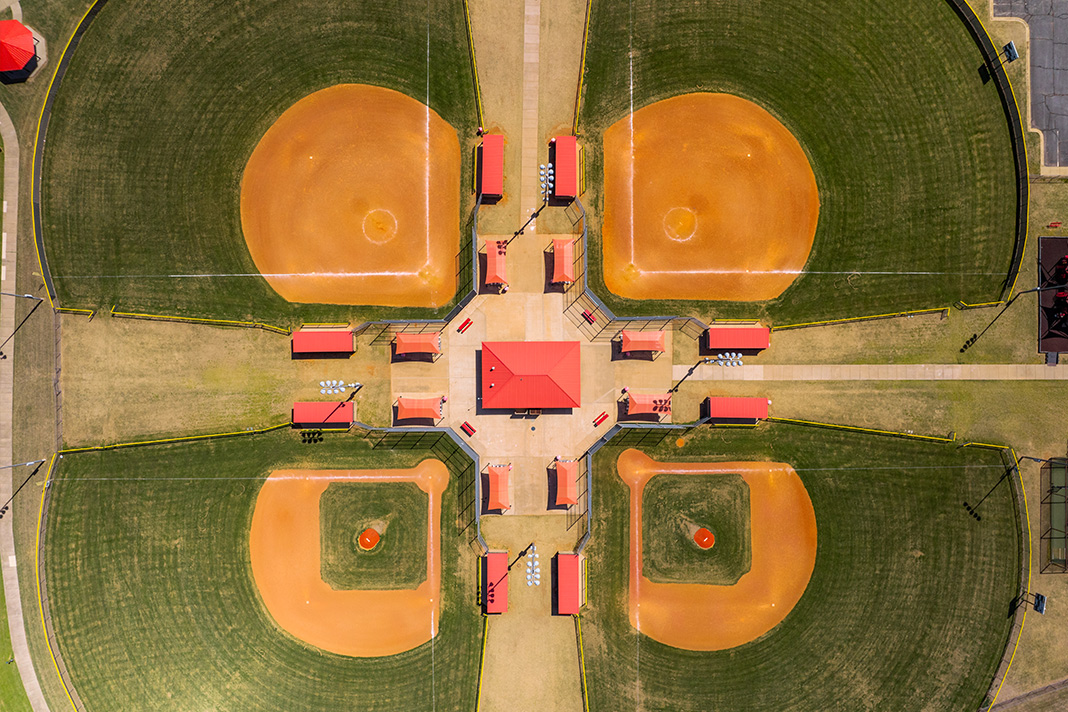  Aerial view of a Farmington symmetrical baseball quad complex with four diamonds, infields, and lush green outfields. Central area with red roofs and seating areas connects all fields